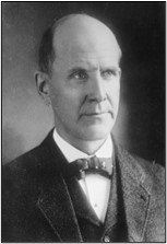A black and white portrait of a man wearing a suit and bowtie, looking directly at the camera.