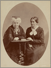 Two women in 19th century attire seated together at a table, with a decorative backdrop.
